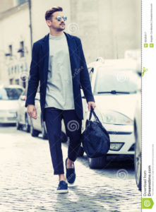 Cool man beautiful model outdoors, city style fashion A handsome man model walking in the city center next to some cars. urban setting. The young boy as trendy, modern clothing with bag. Cobblestones to the ground.