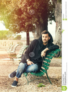 Fashionable cool young man relaxing on a bench in a park with trees A young handsome man sitting on a bench in the historic center of Rome, Italy. The boy dresses fashionable, wearing partially a hoodie. Outdoors in a park.