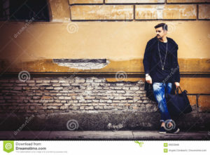 Handsome young man model on street grunge wall Handsome young man model on the go. On the street leaning against the wall. A beautiful model with a black bag is waiting leaning against an ancient wall of an old building.