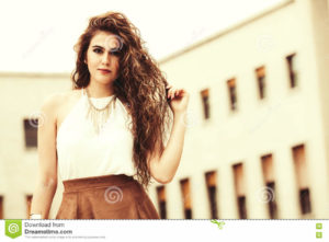 Beautiful and confident woman with curly hair. Urban look A beautiful and confident young woman. Horizontal portrait of a girl with curly hair. Makeup on her face, perfect style. White shirt sleeveless and brown skirt. Sensuality, freshness and beauty. Behind her, a building with windows.