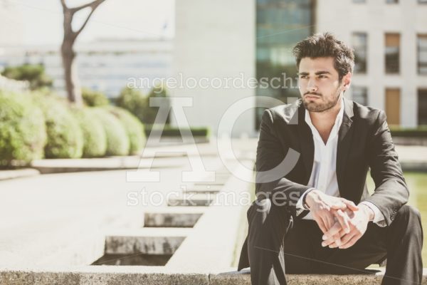 Handsome young man sitting in the city. Relaxed thinking with his arms on her legs. - Angelo Cordeschi